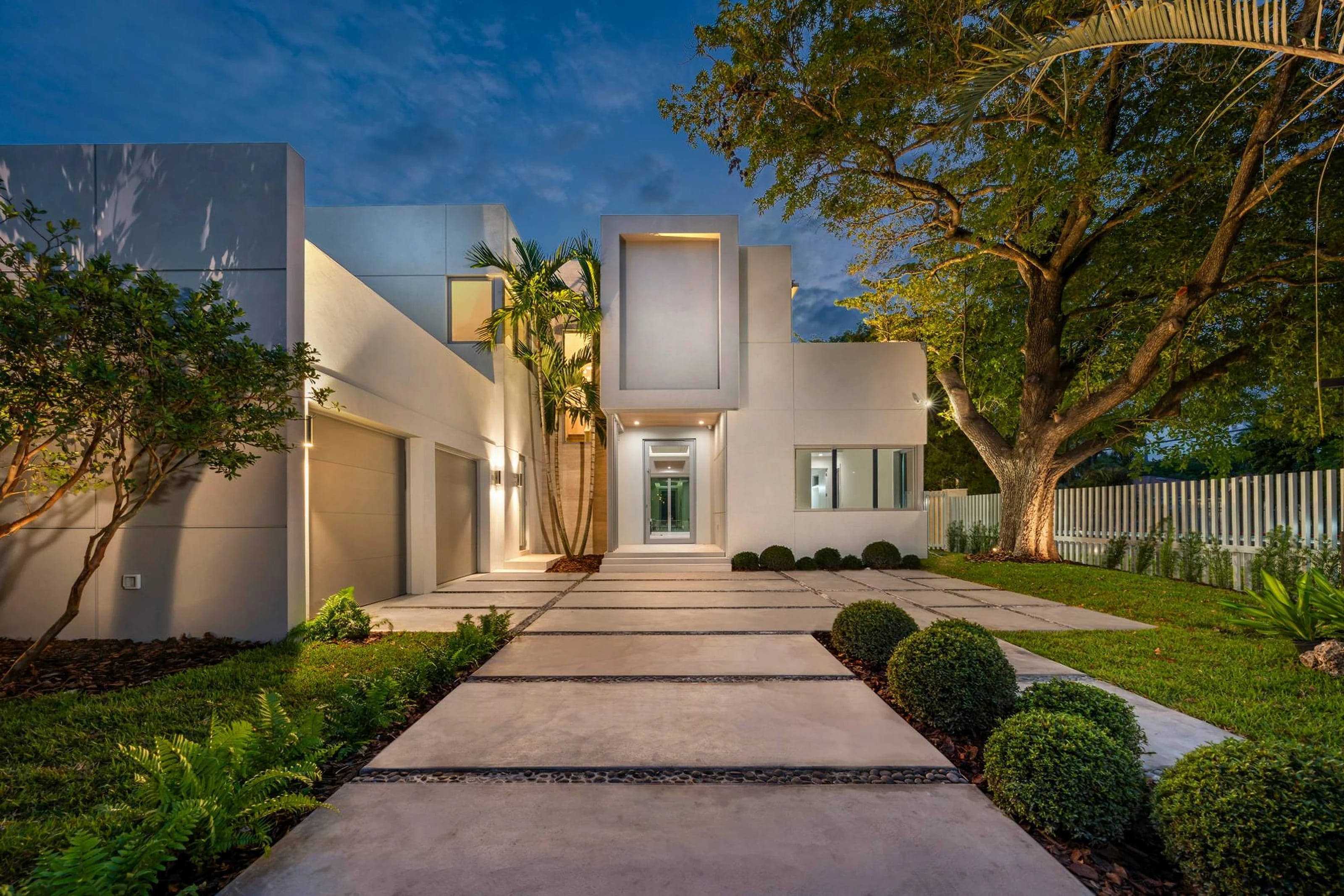 Casa moderna de fachada blanca al atardecer, con camino de losas de hormigón, jardín cuidado y un árbol grande a la derecha
